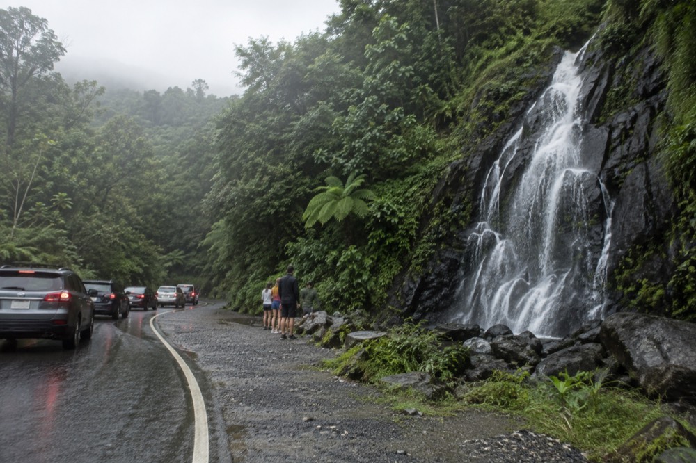 A tall waterfall dropping over a dark rock face beside a paved forest road