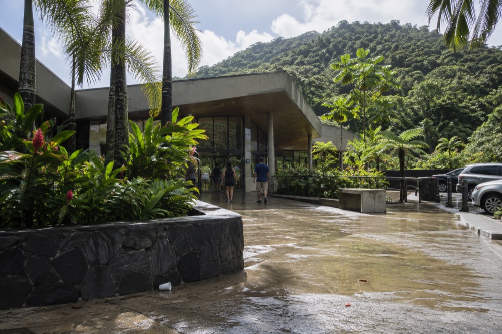 A modern visitor center building with tropical landscaping and a low ridge behind it
