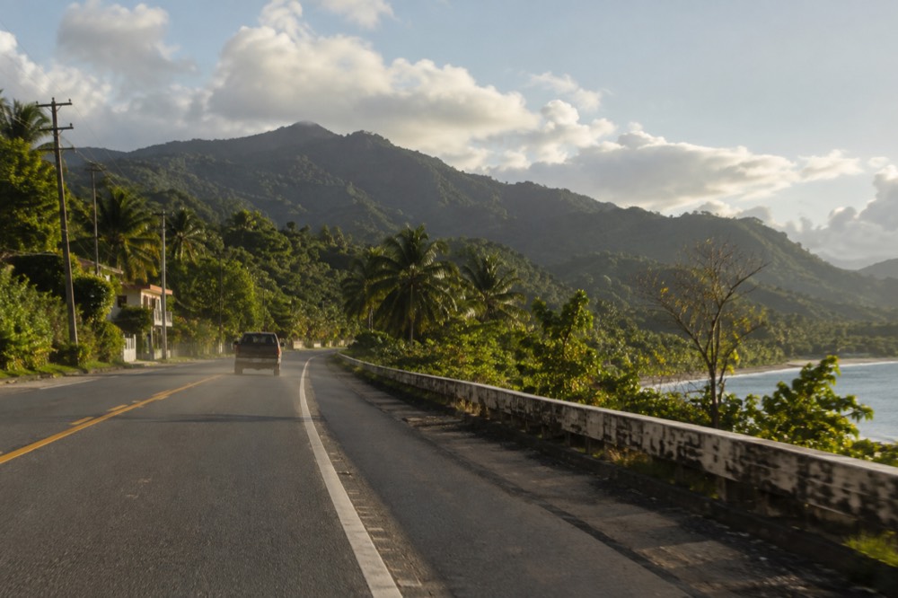 A coastal highway with green mountains rising in the distance under scattered clouds