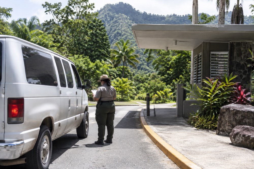 A small passenger van stopped at a signed forest entrance while a ranger checks a clipboard